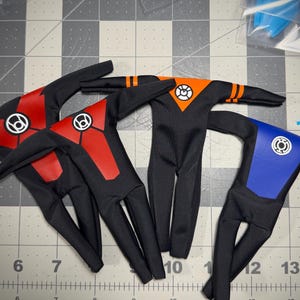 May include: Four miniature black fabric bodysuits for action figures, each featuring a distinct colored chest panel and a circular logo. Two suits have red panels, one has an orange panel with arm stripes, and one has a blue panel.