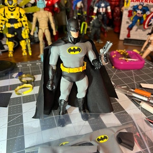 May include: A Batman action figure in a gray and black costume with a yellow utility belt and a black cape. The figure is holding a silver gun. Additional costume pieces are visible. Other superhero figures are in the background.