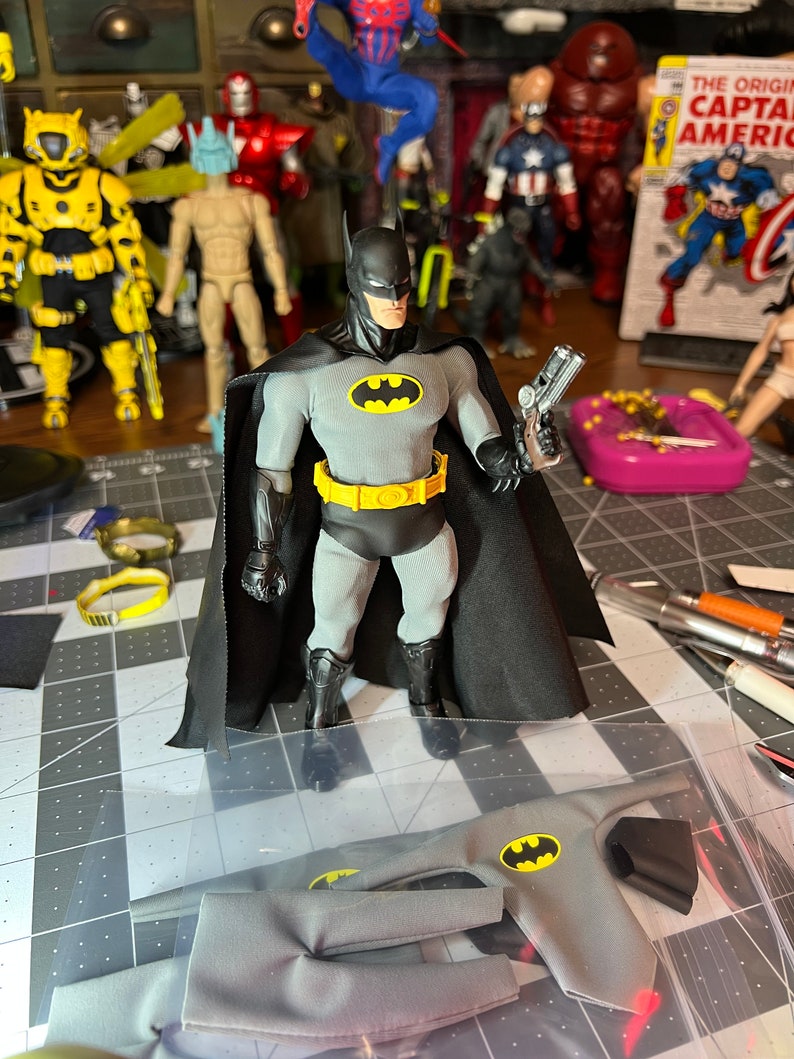 May include: A Batman action figure in a gray and black costume with a yellow utility belt and a black cape. The figure is holding a silver gun. Additional costume pieces are visible. Other superhero figures are in the background.