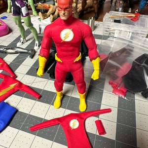 May include: A red and yellow superhero action figure with a lightning bolt symbol on the chest. The figure is standing on a grey and white tiled surface. There are other action figures and accessories in the background.