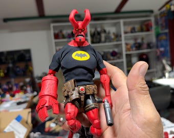 May include: A red Hellboy action figure with two horns, wearing a black t-shirt featuring a yellow bat emblem. The figure has a large red right hand, a brown utility belt with pouches, and holds a small silver-colored cylindrical accessory.