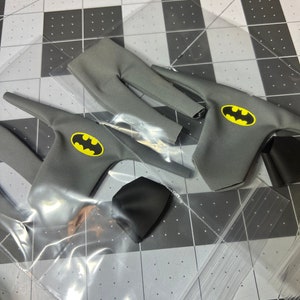 May include: Gray fabric Batman costume pieces, including a top with a yellow bat emblem, pants, and a black cowl. The items are packaged in clear plastic.