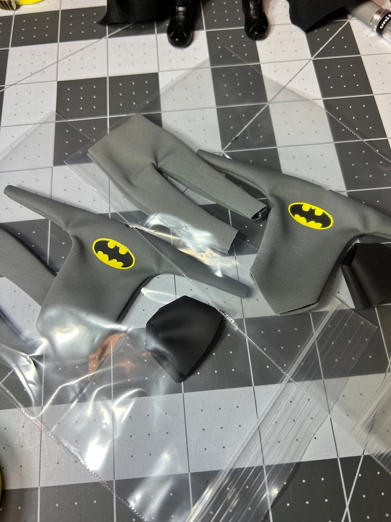 May include: Gray fabric Batman costume pieces, including a top with a yellow bat emblem, pants, and a black cowl. The items are packaged in clear plastic.