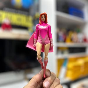 May include: A pink action figure of a female superhero with red hair, wearing a pink bodysuit, cape, and boots. The figure has a symbol on the chest and is held in a hand.
