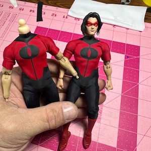 May include: Two action figures, one headless, are displayed. Both figures wear red and black superhero costumes with a circular design on the chest. The figure on the right has black hair and a red mask. The figures are on a pink cutting mat.