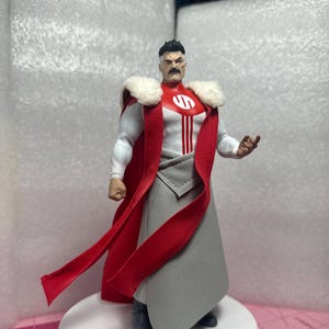 Op de afbeelding: Actiefiguur van een personage in een wit en rood kostuum met een rode cape en grijze rok. De figuur heeft een zwarte snor en haar. De figuur staat op een wit platform.