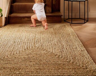 May include: A rectangular jute rug with a woven texture, featuring a geometric design. The rug is a natural beige color and is placed on a wooden floor. A baby in a white onesie is crawling on the rug near a wooden staircase.