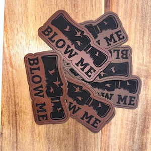 May include: A collection of brown and black leather patches. Each patch features a stylized illustration of a duck call with the words "BLOW ME" in bold letters. The patches are arranged on a wooden surface.