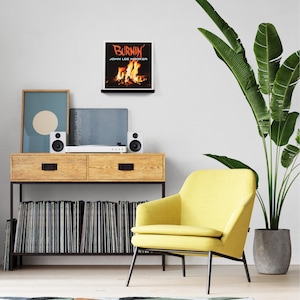 May include: A living room scene featuring a wooden console with a record player, speakers, and a collection of vinyl records. A framed album cover with the text "BURNIN' JOHN LEE HOOKER" hangs above. A yellow armchair and a potted plant complete the decor.