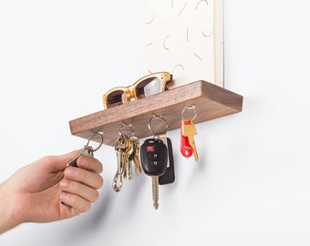 May include: A wooden key rack with five hooks, holding a set of keys and a pair of sunglasses. The rack is mounted on a white wall.