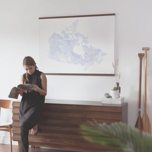 May include: A woman in a black dress reads a magazine next to a wooden dresser. A large framed map of Canada hangs on the wall. Two wooden paddles lean against the wall. A wooden chair is to the left.