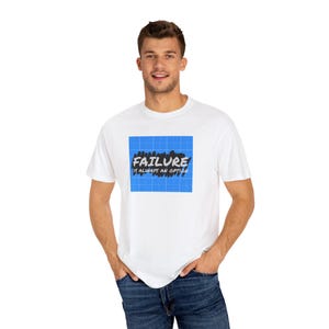 May include: A white short-sleeved t-shirt featuring a central blue square graphic with a white grid pattern. Overlaid on the grid is a black brushstroke shape containing the white text FAILURE IS ALWAYS AN OPTION in a bold, sans-serif font.