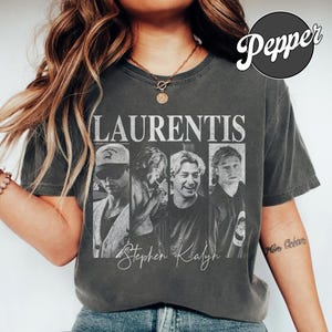 May include: Dark grey t-shirt featuring the word "LAURENTIS" in white above four black and white images of men. The name "Stephen Kalyn" is written in cursive below. The shirt has a vintage look and feel.
