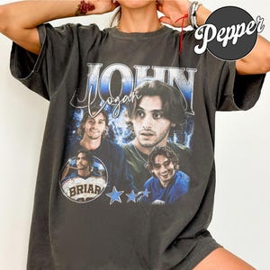 Op de afbeelding: Donkergrijs t-shirt met een collage van afbeeldingen van een man met de naam "John Logan" in een gestileerd lettertype. Het ontwerp bevat blauwe sterren en het woord "Briar" in een cirkel.