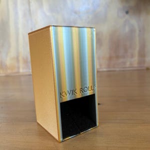 May include: A rectangular dice tower with a textured gold side and a metallic silver front panel featuring vertical yellow stripes. The front is embossed with the text KWIK ROLL and has a black interior opening at the base for dice to exit.
