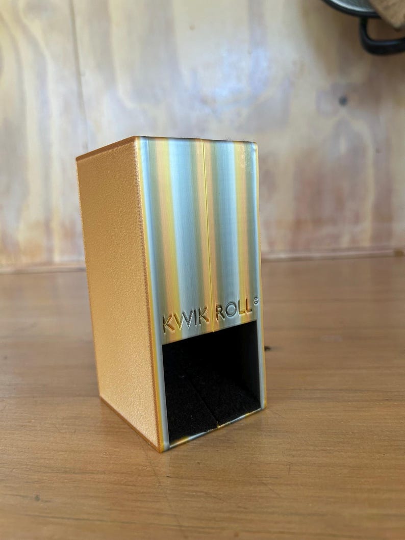 May include: A rectangular dice tower with a textured gold side and a metallic silver front panel featuring vertical yellow stripes. The front is embossed with the text KWIK ROLL and has a black interior opening at the base for dice to exit.