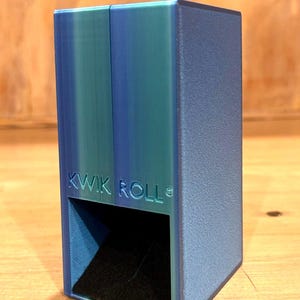 May include: A rectangular blue and teal dice tower with a textured finish. The front face features the embossed text KWIK ROLL above a square opening that reveals a black interior ramp designed for rolling dice during tabletop gaming sessions.