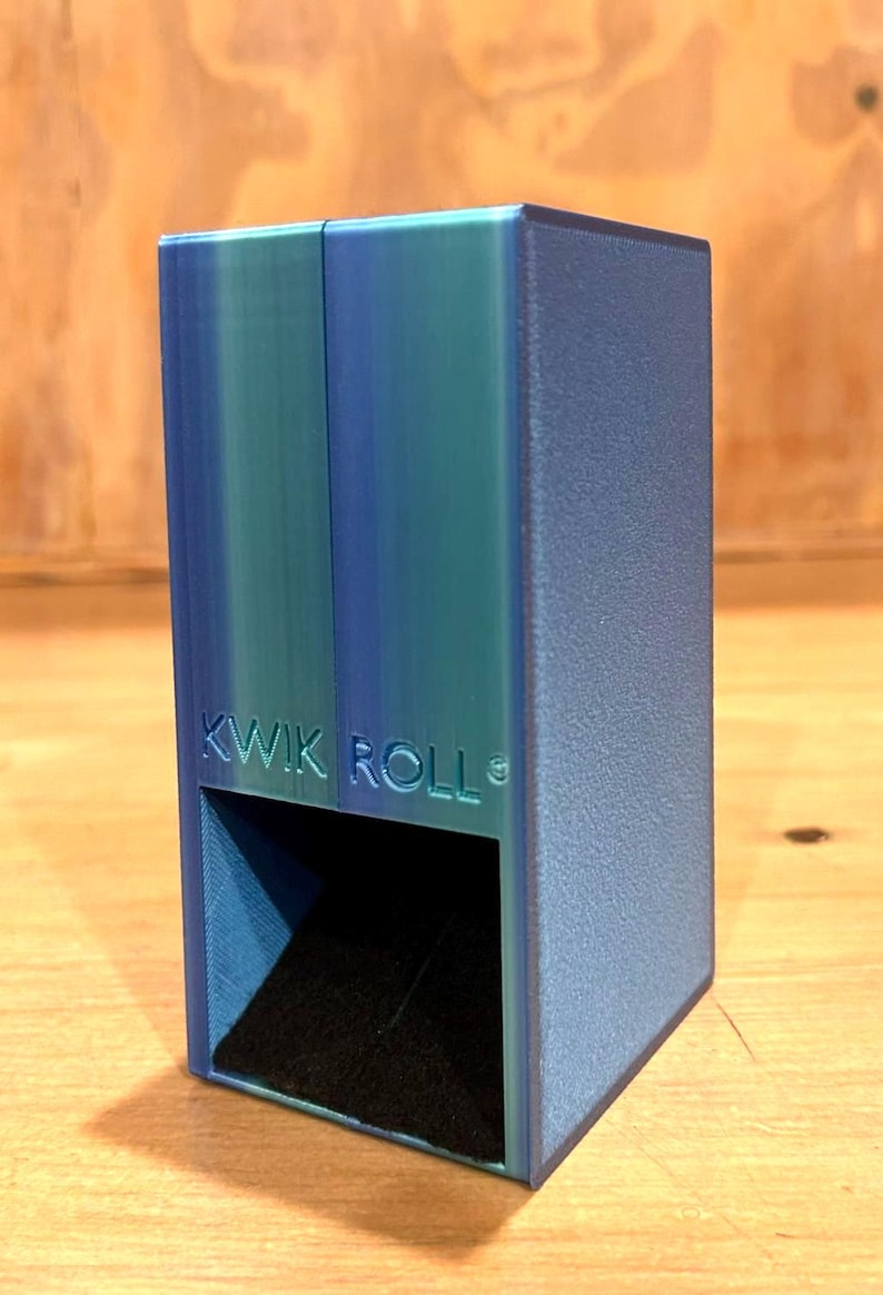 May include: A rectangular blue and teal dice tower with a textured finish. The front face features the embossed text KWIK ROLL above a square opening that reveals a black interior ramp designed for rolling dice during tabletop gaming sessions.