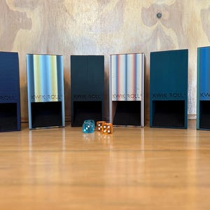 May include: Six rectangular Kwik Roll dice towers in various colors including dark blue, light blue, black, white, teal, and green, each featuring a square opening at the base. Two translucent dice, one blue and one orange, sit in front.