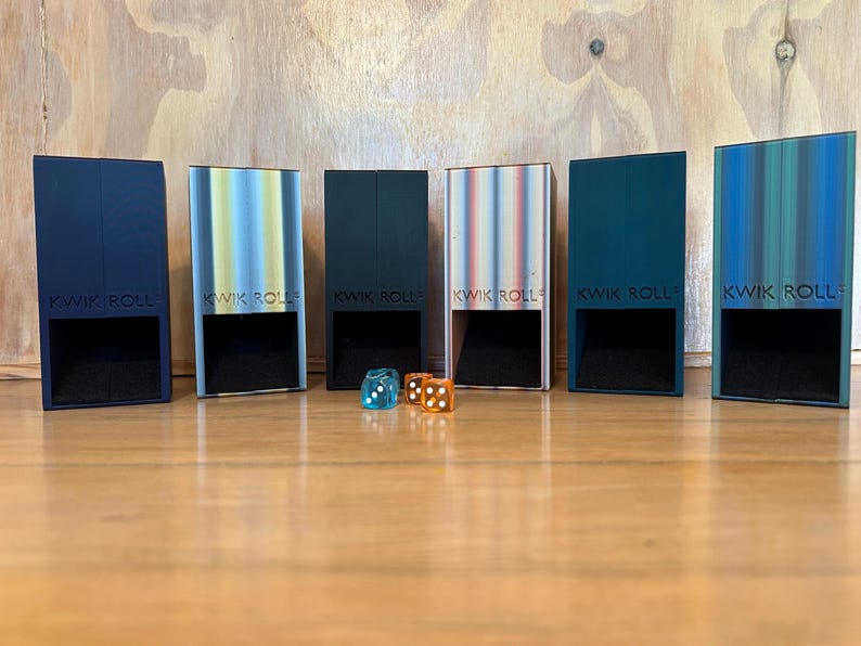 May include: Six rectangular Kwik Roll dice towers in various colors including dark blue, light blue, black, white, teal, and green, each featuring a square opening at the base. Two translucent dice, one blue and one orange, sit in front.