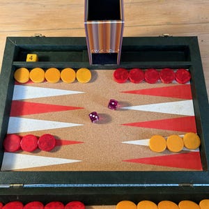 May include: A backgammon board game set featuring a dark green case with a cork playing surface, red and white triangular points, and yellow and red circular checkers. Includes two purple translucent dice and a vertical dice cup labeled KWIK ROLL.