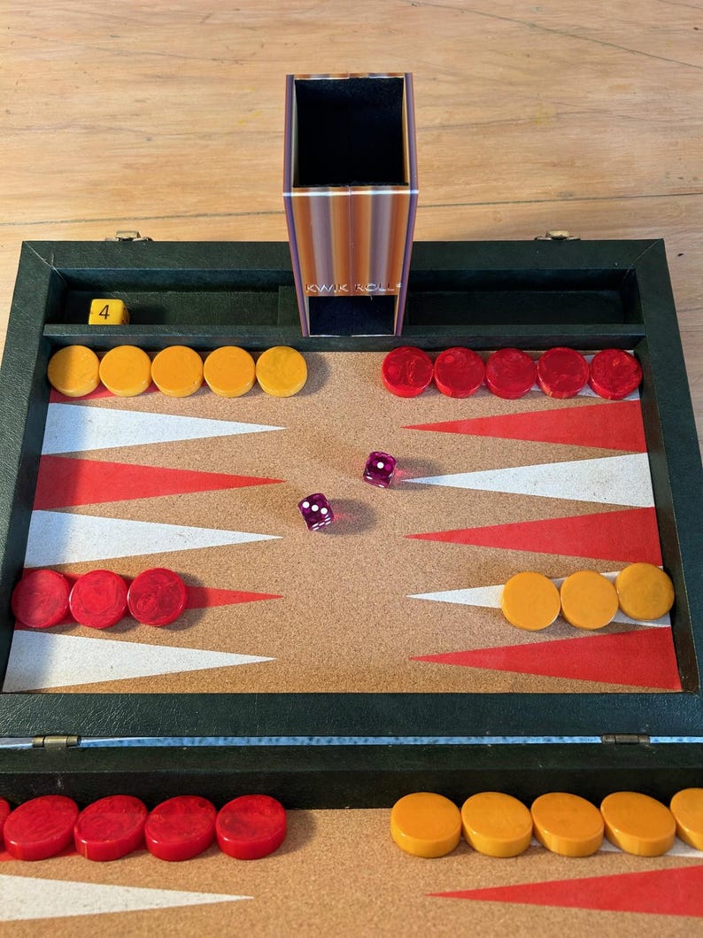 May include: A backgammon board game set featuring a dark green case with a cork playing surface, red and white triangular points, and yellow and red circular checkers. Includes two purple translucent dice and a vertical dice cup labeled KWIK ROLL.