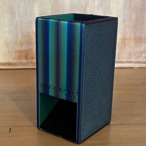 May include: A rectangular dice tower with a textured finish in shades of teal, blue, and dark green. The front features the embossed text KWIK ROLL. It has an open bottom section for dice to exit and a hollow top opening for inserting dice.