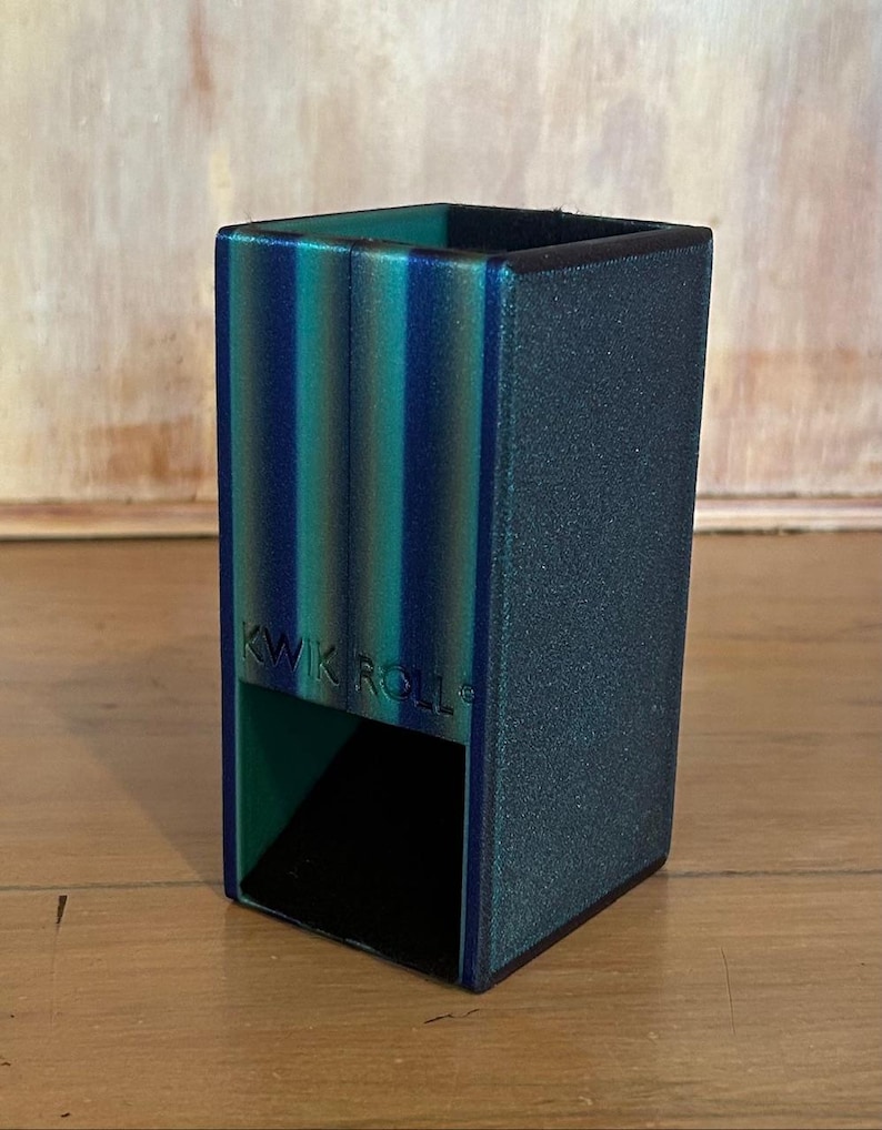 May include: A rectangular dice tower with a textured finish in shades of teal, blue, and dark green. The front features the embossed text KWIK ROLL. It has an open bottom section for dice to exit and a hollow top opening for inserting dice.