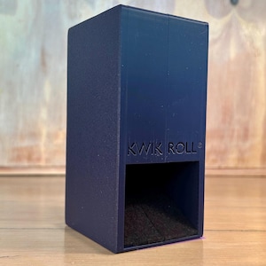 May include: A dark blue rectangular dice tower with a textured finish and the text KWIK ROLL embossed on the front. The device features an open square compartment at the base for dice to exit, with a dark interior surface visible within the opening.