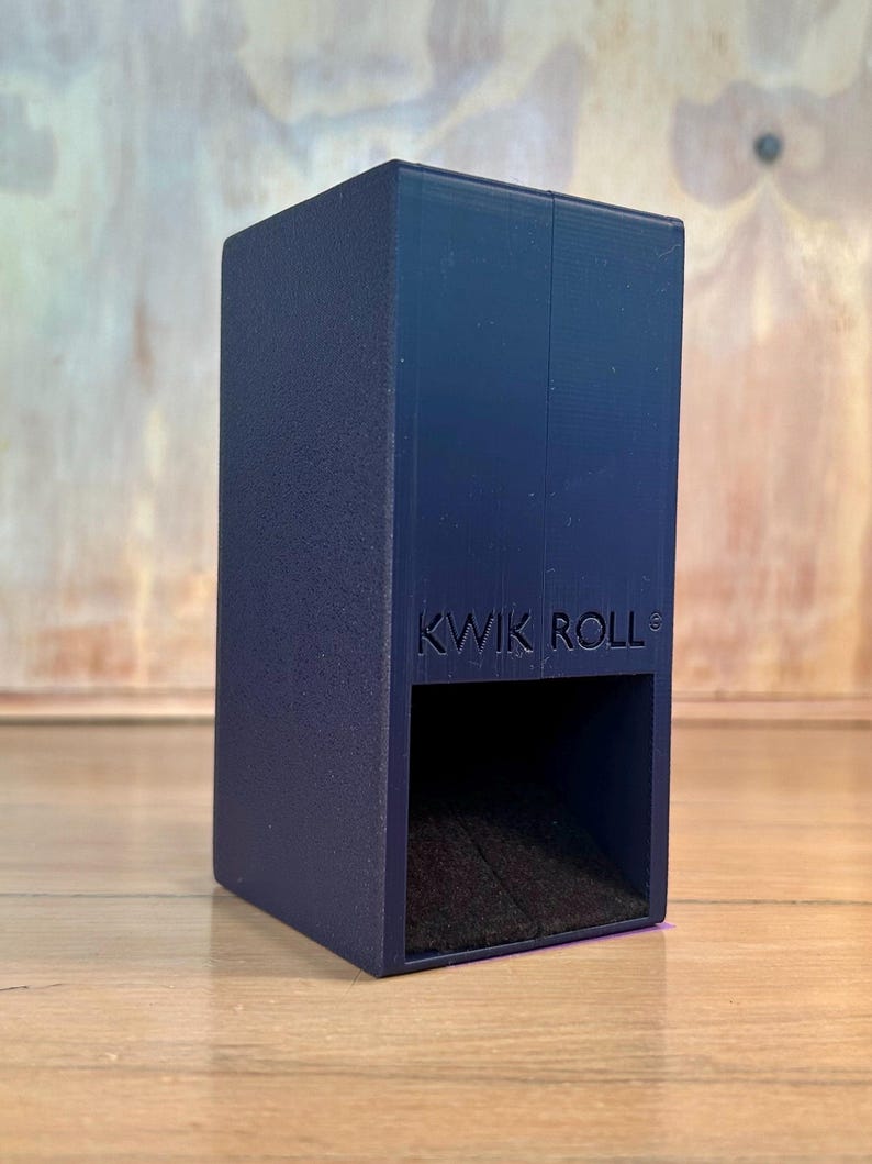 May include: A dark blue rectangular dice tower with a textured finish and the text KWIK ROLL embossed on the front. The device features an open square compartment at the base for dice to exit, with a dark interior surface visible within the opening.