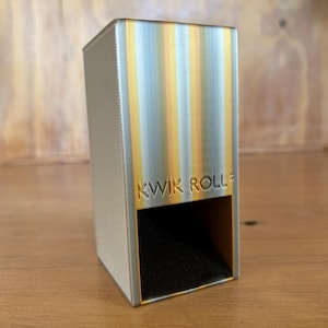 May include: A rectangular Kwik Roll dispenser featuring a metallic silver finish with vertical yellow and orange gradient stripes. The front face displays the embossed text KWIK ROLL above a dark, hollow rectangular opening for dispensing items.