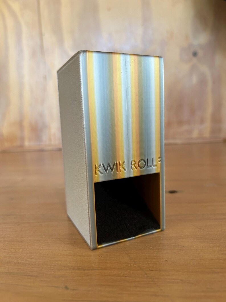 May include: A rectangular Kwik Roll dispenser featuring a metallic silver finish with vertical yellow and orange gradient stripes. The front face displays the embossed text KWIK ROLL above a dark, hollow rectangular opening for dispensing items.