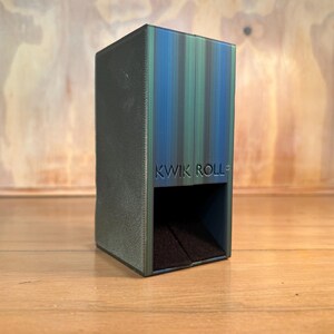 May include: A rectangular dice tower with a gradient finish transitioning from dark green to blue. The front face features the embossed text KWIK ROLL above a square opening that reveals a dark, textured interior base for rolling dice.