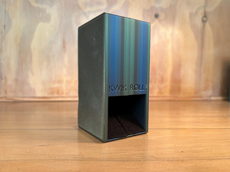 May include: A rectangular dice tower with a gradient finish transitioning from dark green to blue. The front face features the embossed text KWIK ROLL above a square opening that reveals a dark, textured interior base for rolling dice.