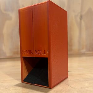 May include: A vertical orange rectangular dice tower with a textured surface and a black felt-lined base. The front of the tower features the embossed text KWIK ROLL with a small copyright symbol. The structure has a hollow opening at the bottom.