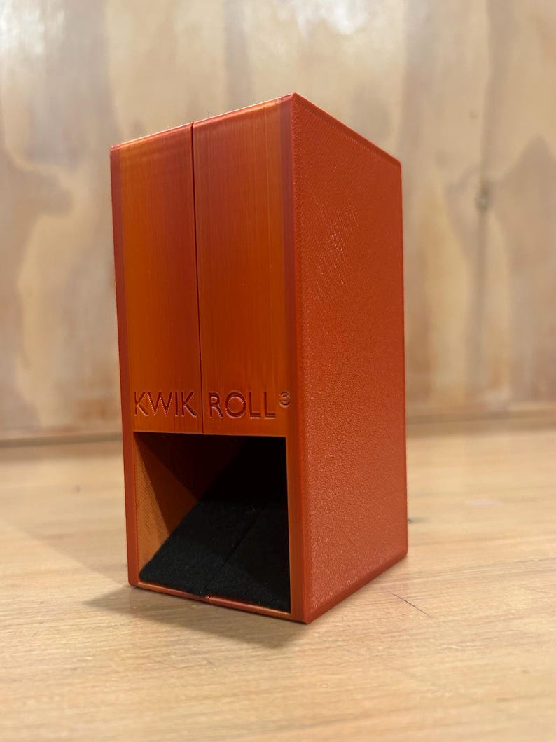 May include: A vertical orange rectangular dice tower with a textured surface and a black felt-lined base. The front of the tower features the embossed text KWIK ROLL with a small copyright symbol. The structure has a hollow opening at the bottom.
