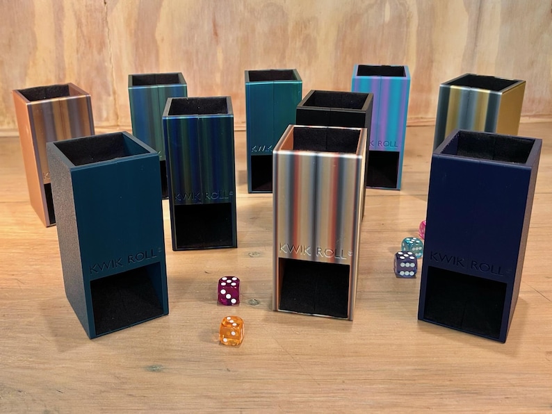 May include: A collection of nine vertical rectangular dice towers in various colors including metallic gradients, solid blue, and black. Each tower features an open base and the embossed text KWIK ROLL on the front. Several multi-colored dice are scattered.