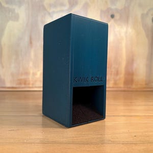 May include: A dark teal, rectangular vertical storage container with the text KWIK ROLL embossed on the front. The object features a hollow, open-faced lower section with a dark interior, designed for holding items in a upright, stable position.