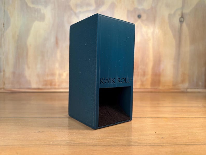May include: A dark teal, rectangular vertical storage container with the text KWIK ROLL embossed on the front. The object features a hollow, open-faced lower section with a dark interior, designed for holding items in a upright, stable position.