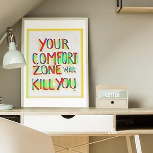 May include: A framed motivational poster with the text "Your comfort zone will kill you" in colorful, rainbow-like lettering. The poster is hanging on a wall above a white desk with a wooden top and white legs. A white chair with wooden legs is in front of the desk.