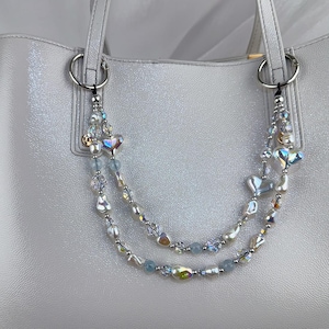 May include: A light gray tote bag with silver hardware and handles. The bag is adorned with two beaded charms featuring iridescent beads, pearls, and heart-shaped accents. The bag has a glossy finish.