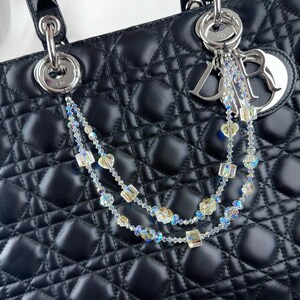 May include: A black quilted leather handbag with silver hardware and a decorative crystal beaded charm. The charm features iridescent beads and a silver logo tag. The bag has a top handle and a diamond pattern.