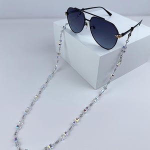 Könnte beinhalten: Schwarze Aviator-Sonnenbrille mit einer silbernen Kette mit schillernden, quadratisch geschnittenen Perlen. Die Sonnenbrille hat einen schwarzen Rahmen mit goldenen Akzenten. Die Kette ist so konzipiert, dass sie die Sonnenbrille um den Hals hält.