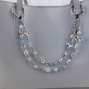 May include: A decorative double-strand bag charm featuring iridescent heart-shaped beads, white baroque pearls, clear faceted crystals, and small light blue round beads. The charm is attached to a light gray handbag with silver-toned metal rings.