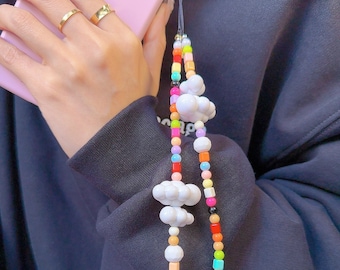 Op de afbeelding: Een kleurrijke kralen telefoonband met witte wolkcharms. De band is gemaakt van kleine, ronde kralen in verschillende kleuren, waaronder rood, oranje, geel, groen, blauw en paars. De band is bevestigd aan een telefoonhoes met een zwart koord.