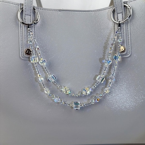 May include: A light gray handbag with silver hardware and a beaded strap. The strap is made of clear, iridescent beads in various shapes, strung on a black cord. The bag has two silver rings for attaching the strap.