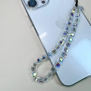 May include: A white smartphone with a triple-lens camera system features a decorative phone charm attached to its side. The charm consists of a black cord holding a series of clear, square-shaped beads that display iridescent, rainbow-like reflections.