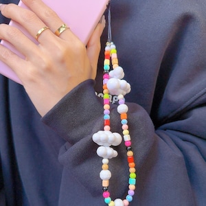 Puede incluir: Una correa de teléfono con cuentas de colores y dijes de nubes blancas. La correa está hecha de pequeñas cuentas redondas de varios colores, incluyendo rojo, naranja, amarillo, verde, azul y morado. La correa está unida a una funda de teléfono con un cordón negro.