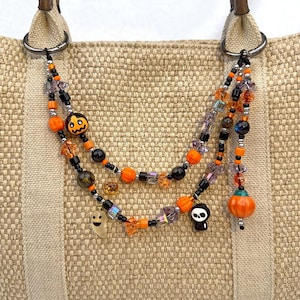 May include: A multi-strand beaded bag charm featuring orange, black, and purple beads. The accessory includes charms shaped like a jack-o'-lantern, a ghost, a skull, and a pumpkin, attached to the metal rings of a beige woven tote bag.
