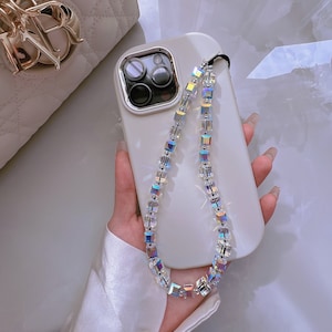 Puede incluir: Una funda de teléfono blanca con una correa de teléfono de cristal iridiscente transparente. La correa está hecha de cristales de forma cuadrada.